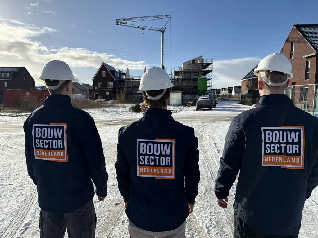 Drie vakmensen van Bouwsector Nederland met witte helm en donkerblauwe jas, van achteren, kijken naar een woonwijk in aanbouw